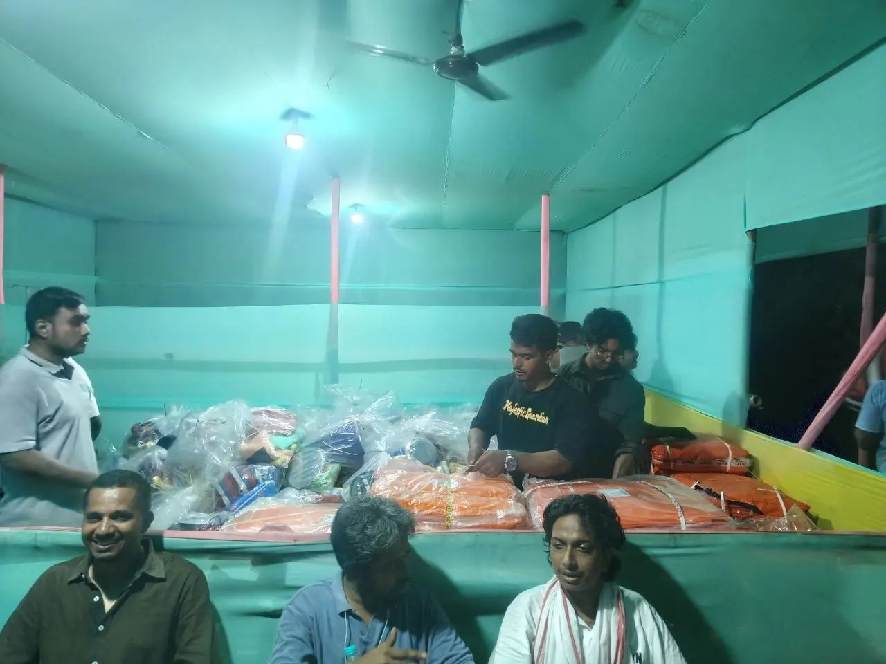Assam, August 2026: Humrahi volunteers handing wrapped relief kits across a bench to seated recipients during the night distribution at Mautgaon