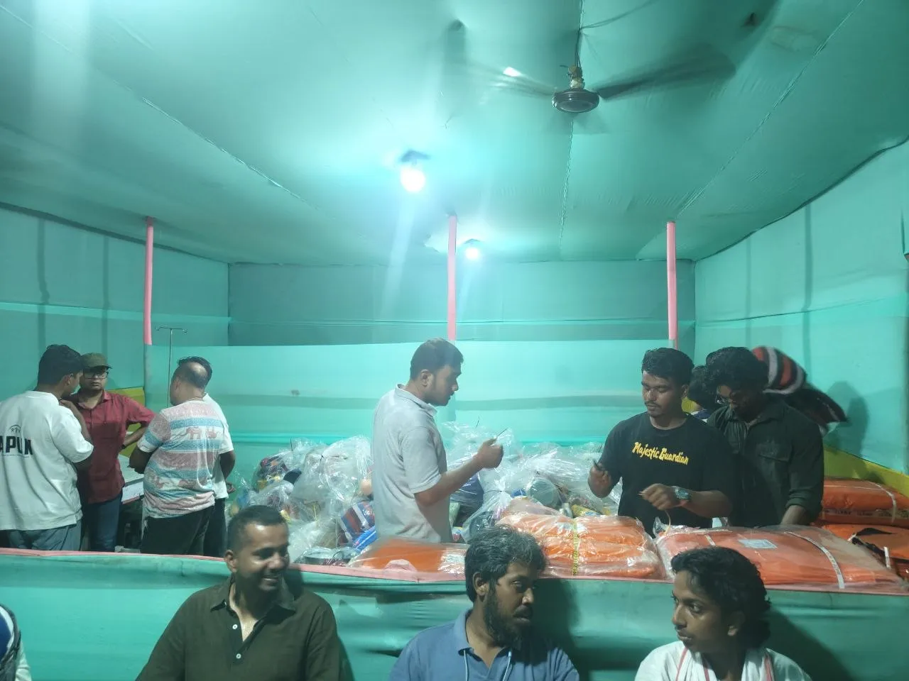 Volunteers sorting wrapped relief supplies on a table inside the shelter hall at night, men seated in the foreground