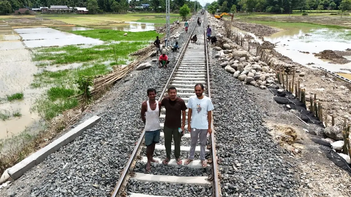 Three residents standing on a raised railway line in flood-hit Assam, the ballast washed out and packed back with bamboo piling, flooded paddy fields on both sides
