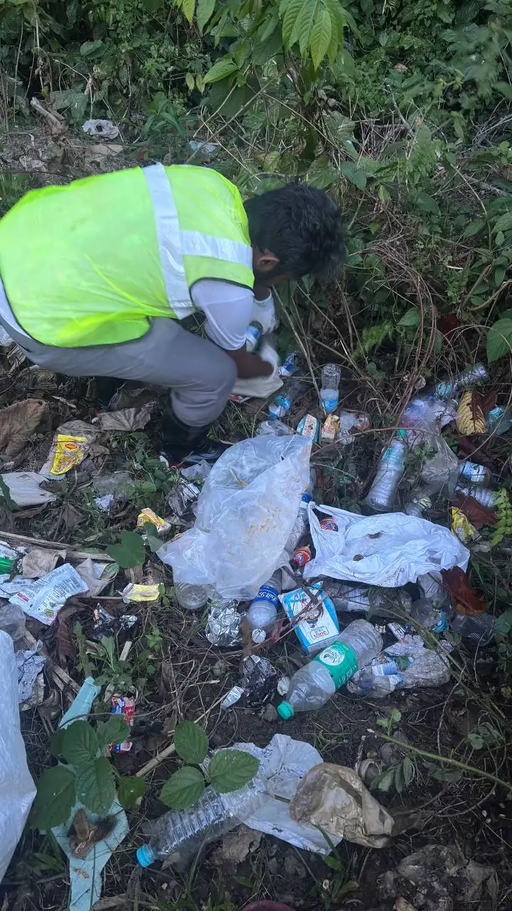 A volunteer in a hi-vis vest crouched among dense plastic bottles and wrappers on the roadside verge