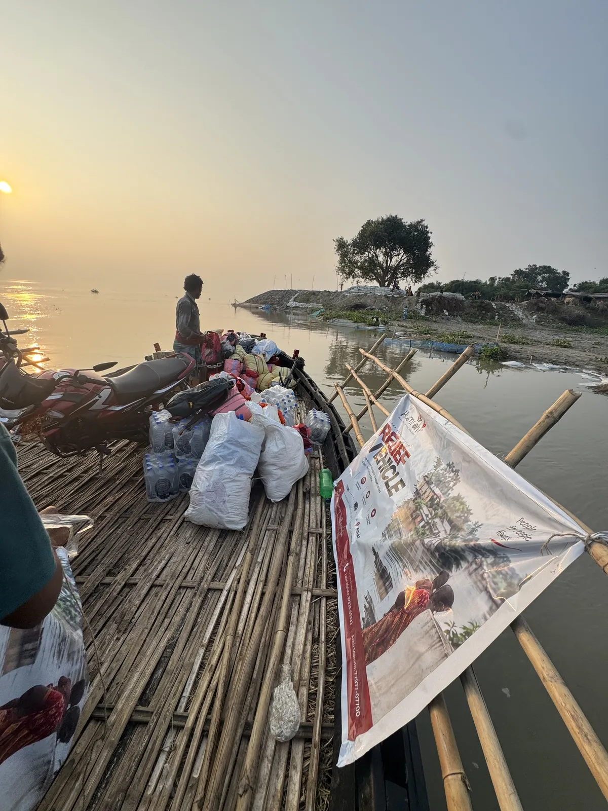 Bihar, 19 September 2026: relief sacks and bottled water stacked on the bamboo deck of a country boat, the Relief Vehicle banner tied along its side