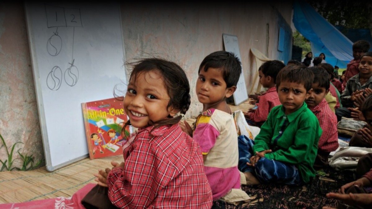 Children sitting on mats with a storybook propped against a whiteboard, at a Support Wings Shiksha Sabka Haq session. This is not Siliguri and not a children's home library.